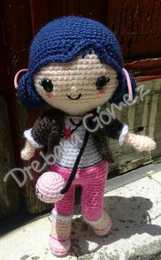 a crocheted doll with blue hair and pink pants is standing on the sidewalk