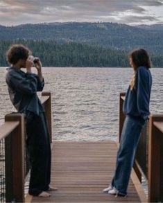 two people are standing on a dock looking at the water and mountains in the distance