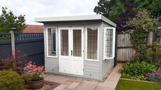 a small gray shed with white doors and windows in the back yard next to a flower garden