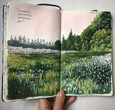 a hand holding an open book with watercolors on the pages and trees in the background