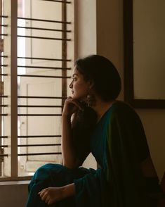 a woman sitting in front of a window with her hand on her chin looking out the window