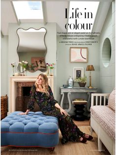 a woman sitting on top of a blue ottoman in front of a mirror and fireplace