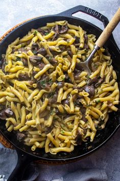 a skillet filled with pasta and mushrooms on top of a table next to a wooden spoon