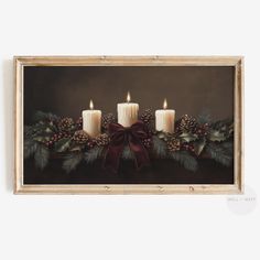 three lit candles with holly and pine cones in front of a brown background, framed on the wall