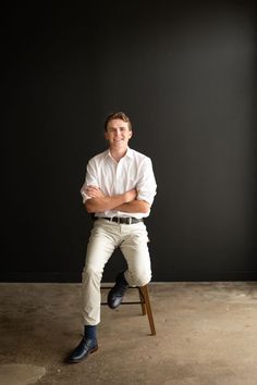 a man sitting on top of a wooden chair in front of a black wall with his arms crossed