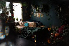 a bed with blue sheets and pillows in a dark room next to a window filled with potted plants