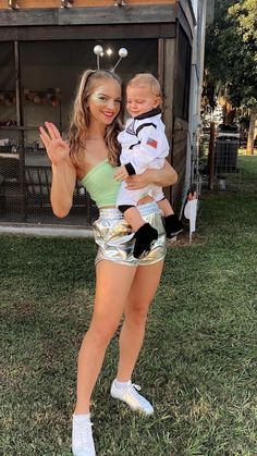 a woman holding a baby in her arms while standing on the grass with another person behind her