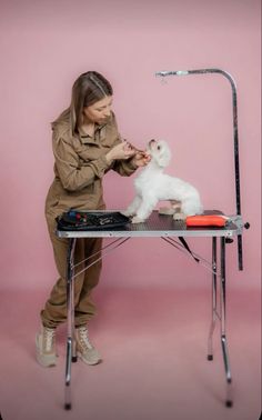 a woman grooming a small white dog on top of a table with a pink background