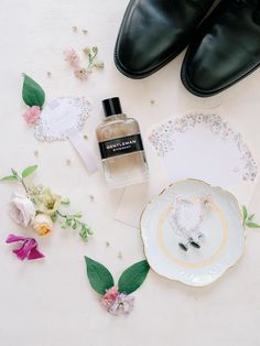 a pair of black shoes next to a bottle of perfume and some flowers on a white surface