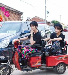 two people riding on the back of a small red scooter next to a black car