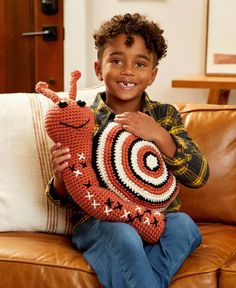 a young boy sitting on a couch holding a crocheted snail pillow and smiling at the camera