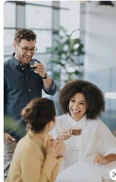 two women and a man having drinks at a table in an office with other people