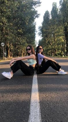 two young women sitting on the side of an empty road with trees in the background