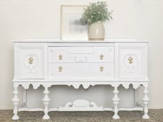 an ornate white dresser with gold details and a potted plant on top, against a wall