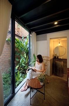 a woman sitting on a chair reading a book in front of a large window with plants outside