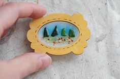 a hand holding a small wooden brooch with three green trees in the center and red berries on it