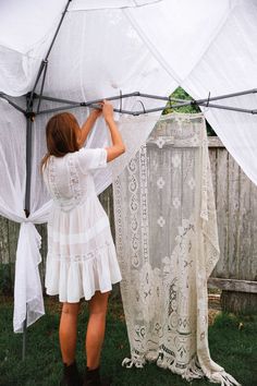 a woman in white dress standing next to an open tent with curtains hanging from it