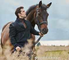 a man standing next to a brown horse on top of a lush green field with tall grass