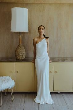 a woman in a white dress standing next to a table with a lamp on it