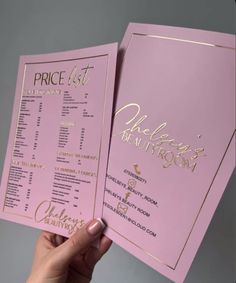 a person holding up two pink menus with gold lettering on them and the price list