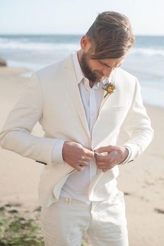 a man in a white suit is looking down at his watch while standing on the beach