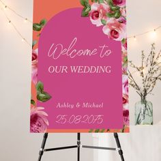 a welcome sign with pink flowers on an easel in front of a string of lights