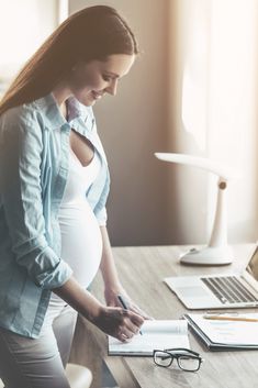 a pregnant woman sitting at a desk in front of a laptop and writing on a notepad