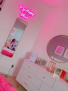 a bedroom with pink walls and white dressers in the corner, there is a neon sign on the wall above the mirror