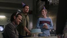 three men standing around a table with a cake on it and one man holding a cup