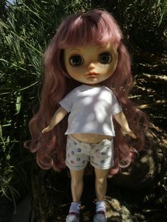 a doll with pink hair standing in front of some bushes and grass, wearing shorts