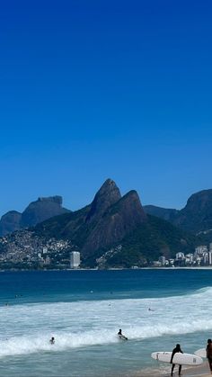 several people are walking on the beach with surfboards in hand and mountains in the background