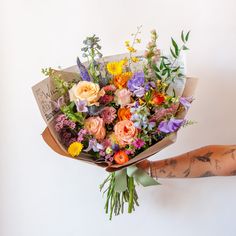 A tattooed arm holds a bouquet wrapped in floral newspaper and brown kraft paper in front of a white backdrop. It's filled with pink, orange, yellow, and purple flowers, including piccolini daisies, roses, veronica, sweet pea, clematis, delphinium, oncidium orchid, and lisianthus. Wildflower Bridal Bouquets, Wildflower Wedding Theme, Wildflower Wedding Bouquet, Summer Wedding Bouquets, Boquette Flowers, Spring Wedding Flowers, Colorful Bouquet, Wildflower Bouquet, Wildflower Wedding