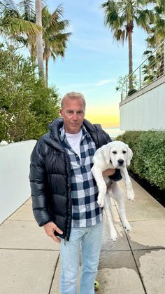 a man holding a white dog in his arms on a sidewalk with palm trees behind him