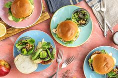 three plates with burgers on them and silverware next to an apple, orange juice, and other food items