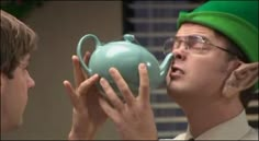 a man in a green hat is holding a teapot to another man's face