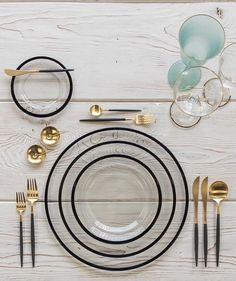 a table setting with black and gold plates, silverware, and glasses on a white wooden surface