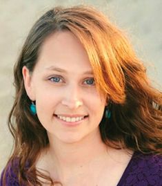 a woman with red hair and blue earrings smiling at the camera while wearing a purple shirt