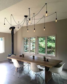 a dining room table with white chairs and lights hanging from the ceiling above it in front of two windows