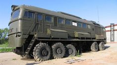 an army truck with large tires parked on the side of a dirt road in front of a building