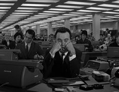 a man sitting at a desk in an office surrounded by people working on computers and telephones