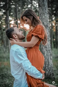 a pregnant woman in an orange dress is holding her husband's back as they stand in the woods