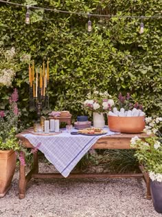 a table with flowers and candles on it in front of a bushy wall, surrounded by potted plants