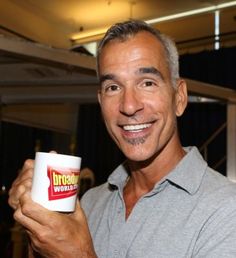 a man holding a coffee cup in his right hand and smiling at the camera while wearing a gray shirt