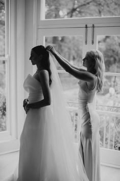 two women standing next to each other in front of a window wearing wedding gowns