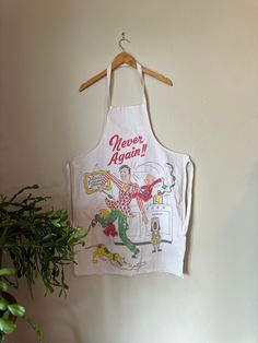 an apron hanging on a wall with a plant in front of it and a sign that says never again