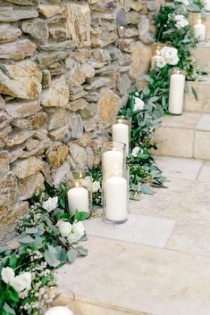 candles are lined up on the side of a stone wall with greenery and flowers