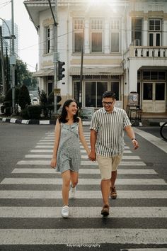 a man and woman walking across a cross walk holding hands in front of a building