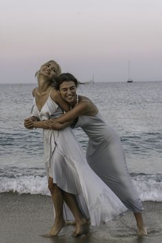 two women hugging on the beach with boats in the water and one woman is wearing a gray dress