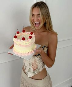 a woman holding a cake with cherries on it in her right hand and smiling at the camera