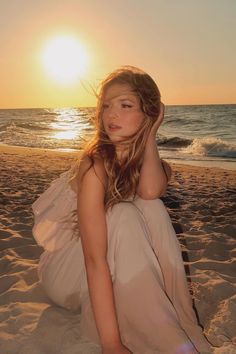 a woman sitting on top of a sandy beach next to the ocean at sun set